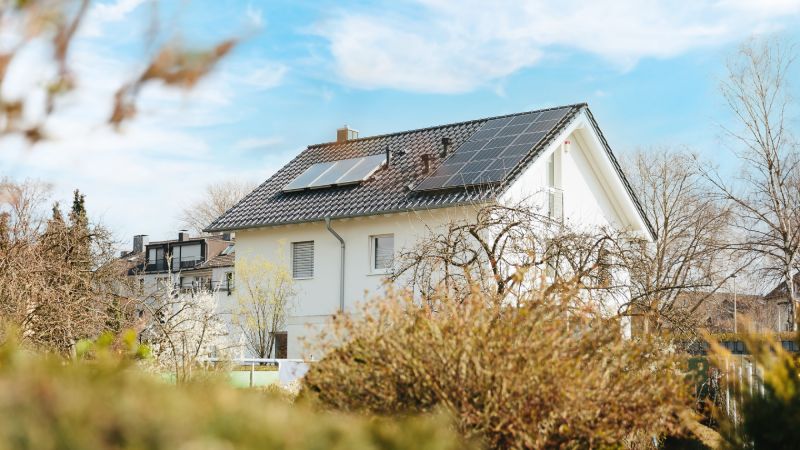 Haus mit Photovoltaikanlage auf dem Dach hinter Sträuchern und Bäumen